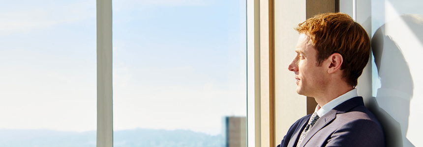 A businessman stands against a wall, looking out a large office window.